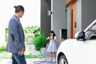 Progressive father and daughter plugs EV charger from home charging station to electric vehicle. Future eco-friendly car with EV cars powered by renewable source of clean energy.