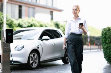 İş kadını tablet kullanıyor, halka açık otoparkta elektrikli aracını şarj ederken yürüyor. Teknolojinin ilerici yaşam tarzı ve EV otomobilinin ekolojik kaygısı