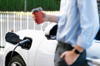 Progressive eco-friendly concept of focus parking EV car at public electric-powered charging station in city with blur background of businessman waiting on recharging car with coffee.