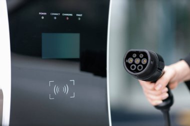Focus closeup hand holding EV plug for electric vehicle pointing at camera with charging station at background as progressive idea of alternative sustainable clean energy for ecological awareness idea