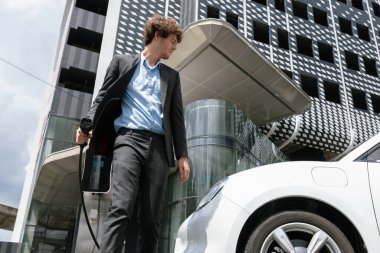 Below view of progressive businessman with electric car recharging at public charging station at modern city residential building. Eco friendly rechargeable EV car powered by alternative clean energy.