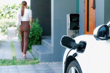 Focus EV charger plugged into EV car at home charging station with blurred background of progressive woman walking in background. Elective vehicle powered by clean energy for eco-friendly concept.