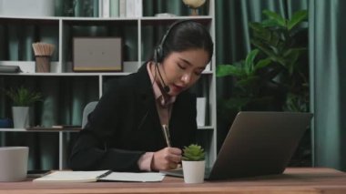 Kulaklık ve mikrofon takan iş kadını masasında çalışıyor ve dizüstü bilgisayar kullanıyor. Uzaktan kumandalı online toplantı için dizüstü bilgisayar kullanan hevesli kadın çalışanlar, ofiste iş videosu konferansı.