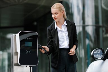 Progressive businesswoman insert charger plug from charging station to her electric vehicle with apartment condo building in background. Eco friendly rechargeable car powered by sustainable energy.