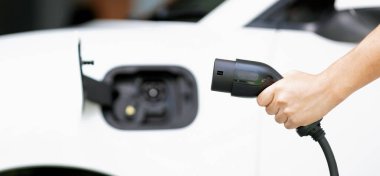 Focus hand holding EV charger plug with blurred background of progressive electric vehicle and socket parking in home garage with electric charging station powered by clean and sustainable energy.