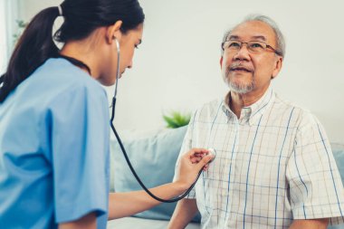 Caring young female doctor examining her contented senior patient with stethoscope in living room. Medical service for elderly, elderly sickness, declining health.