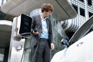 Below view of progressive businessman with electric car recharging at public charging station at modern city residential building. Eco friendly rechargeable EV car powered by alternative clean energy.