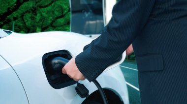 İlerici işadamı elektrikli aracını şarj kablosu ile şarj ediyor. Şarj istasyonundan şehrin arka planına bakıyor. Modern temiz enerji kampanyası konsepti için..