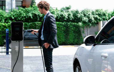 Modern şehir yerleşim bölgesindeki kamu şarj istasyonunda elektrikli araba şarj eden yenilikçi bir işadamı. Çevre dostu şarj edilebilir araba alternatif temiz enerjiyle çalışıyor..