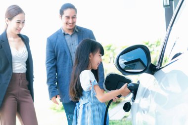 Focus closeup electric vehicle recharging battery from home electric charging station with blurred family in background. Renewable clean energy car for progressive eco awareness lifestyle concept.