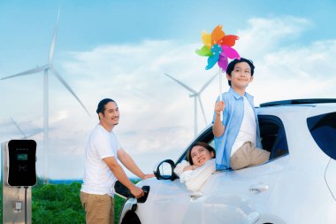 Concept of progressive happy family holding windmill toy and relax at wind farm with electric vehicle. Electric vehicle driven by clean renewable energy from wind turbine generator to charger station.