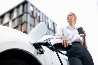 Şehir merkezinde dolaşmadan önce elektrikli arabası ve şarj istasyonu olan ilerici bir iş kadını. Çevre dostu şarj edilebilir EV araba sürdürülebilir ve temiz enerjiyle çalışıyor.