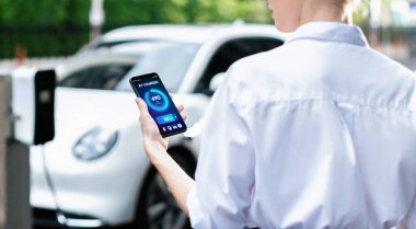 Suit-clad progressive businesswoman look at EV cars battery status from her phone while standing on a charging station with a power cable plug and a renewable energy-powered electric vehicle.