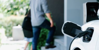 Arka planda bulanık baba ve kızının yürüdüğü şarj istasyonunda elektrikli araba şarj etmeye odaklan. Sağlıklı çevre yaşam tarzı konsepti için yenilikçi yeşil ve temiz enerji aracı.