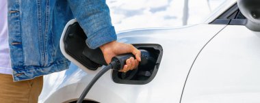 Elektrikli arabasıyla ilerici bir adam, EV arabası gelecekteki sürdürülebilir enerji kavramı olarak rüzgar türbiniyle yeşil alandaki enerji şarj etme istasyonundan enerji yüklüyor. Enerji üreteci olan elektrikli bir araç..