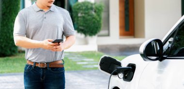 Progressive asian man install cable plug to his electric car with home charging station in the backyard. Concept use of electric vehicles in a progressive lifestyle contributes to clean environment.