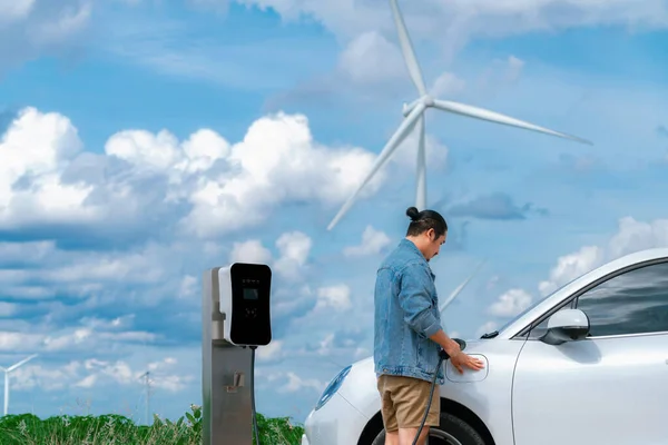 电动汽车是一个进步的人 他的电动汽车 电动汽车用风力涡轮机作为未来可持续能源的概念 从绿地的充电站向能源充电 配备发电机的电动车 — 图库照片