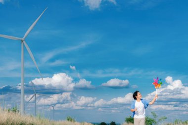 Progressive young asian boy playing with wind pinwheel toy in the wind turbine farm, green field over the hill. Green energy from renewable electric wind generator. Windmill in the countryside concept