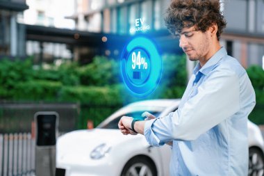 Progressive businessman check wearable hologram watch on electric car battery that being recharge at charging station point in the city. Digital interface, data from EV car display on smartwatch. clipart