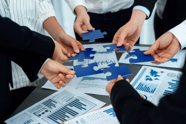 Closeup top view business team of office worker putting jigsaw puzzle together over table filled with financial report paper in workplace with manager to promote harmony concept in meeting room.
