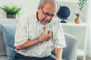 An agonizing senior man suffering from chest pain or heart attack alone in his living room. Serious health problem and feeling unwell concept for seniors.