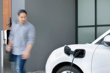 Focus electric car charging at home charging station with blurred progressive man walking in the background. Electric car using renewable clean for eco-friendly concept.