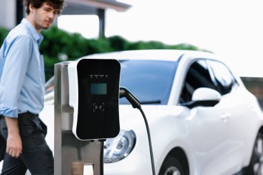 Progressive eco-friendly concept of focus parking EV car at public electric-powered charging station in city center with blur background of businessman waiting at his recharging-electric vehicle.