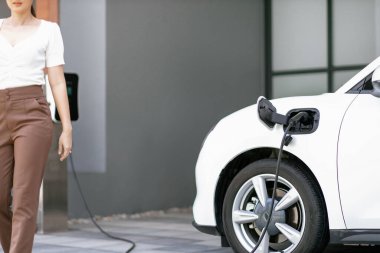 Focus image of electric vehicle recharging battery at home charging station with blurred woman walking in the background. Progressive concept of green energy technology applied in daily lifestyle.