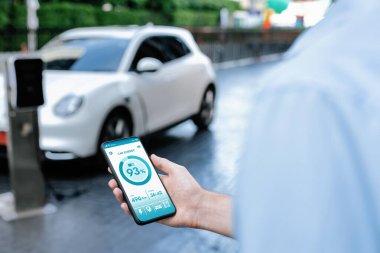Closeup progressive businessman look at EV cars battery status application on smart phone screen at public parking charging station with power cable plug and renewable energy-powered electric vehicle