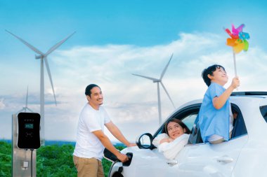 Concept of progressive happy family holding windmill toy and relax at wind farm with electric vehicle. Electric vehicle driven by clean renewable energy from wind turbine generator to charger station.