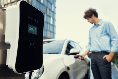 Focus public EV charging station in city residential area with blur background of businessman charging electric vehicle. Progressive lifestyle of ecological concern of city-dwelling individual.
