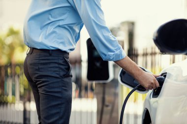 Closeup progressive man holding EV charger plug from public charging station for electric vehicle with background of residential building as concept eco-friendly sustainability energy car concept.
