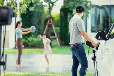 Focus image of progressive man charging electric car from home charging station with blur mother and daughter playing together in the background.