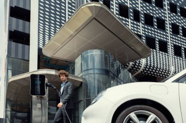 Below view of progressive businessman with electric car recharging at public charging station at modern city residential building. Eco friendly rechargeable EV car powered by alternative clean energy.