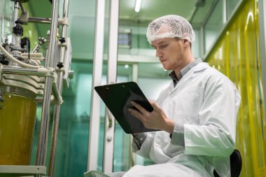 Eczacı bilim adamı bir CBD yağ çıkarıcısından bilgi almak için bir pano ve kalem kullanıyor ve tıbbi esrar ürünleri üretmek için bilimsel bir makine kullanıyor..