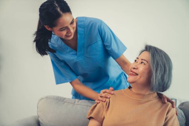 A caregiver rest her hands on the shoulders of a contented senior patient while she sitting on the sofa at home.