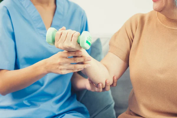 Caregiver helping contented senior woman exercise with dumbbell at home. Rehab for elder, senior medical care.