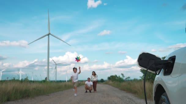 電気自動車で風力発電所での時間を楽しむ進歩的な幸せな家族の概念 風力発電機からの再生可能エネルギーを利用した充電ステーション用電気自動車 — ストック動画
