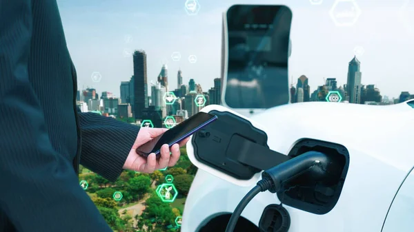 Progressive green city ESG symbol background with electric vehicle. Businessman checking EV cars battery with smartphone at charging station in the urban with ecological and green symbols signs.