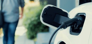 Focus electric car charging at home charging station with blurred progressive man walking in the background. Electric car using renewable clean for eco-friendly concept.