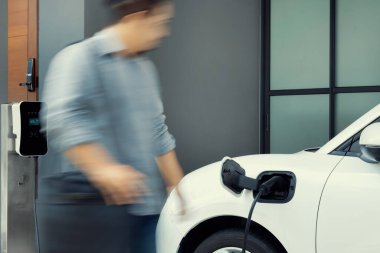 Focus electric car charging at home charging station with blurred progressive man walking in the background. Electric car using renewable clean for eco-friendly concept.
