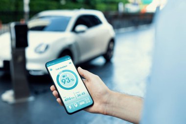 Closeup progressive businessman look at EV cars battery status application on smart phone screen at public parking charging station with power cable plug and renewable energy-powered electric vehicle