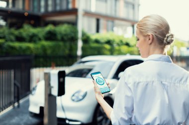 Suit-clad progressive businesswoman look at EV cars battery status from her phone while standing on a charging station with a power cable plug and a renewable energy-powered electric vehicle.
