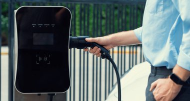 Closeup progressive man holding EV charger plug from public charging station for electric vehicle with background of residential building as concept eco-friendly sustainability energy car concept.