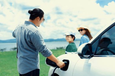 Concept of progressive happy family enjoying their time at green field and lake with electric vehicle. Electric vehicle driven by clean renewable from eco-friendly power sauce.