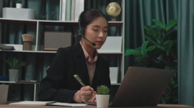 Kulaklık ve mikrofon takan iş kadını masasında çalışıyor ve dizüstü bilgisayar kullanıyor. Uzaktan kumandalı online toplantı için dizüstü bilgisayar kullanan hevesli kadın çalışanlar, ofiste iş videosu konferansı.
