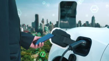 İlerleyen yeşil şehir ESG sembolü, elektrikli araçla birlikte. İş adamı EV arabalarını şarj etmek için şarj aletini şarj ediyor. Şehirdeki şarj istasyonunda ekolojik ve yeşil semboller var..