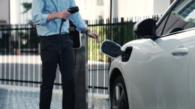 İlerici işadamı, şehir merkezinde dolaşmadan önce şarj cihazından elektrikli arabasına şarj prizi takıyor. Çevre dostu şarj edilebilir araba, sürdürülebilir ve temiz enerjiyle çalışıyor..