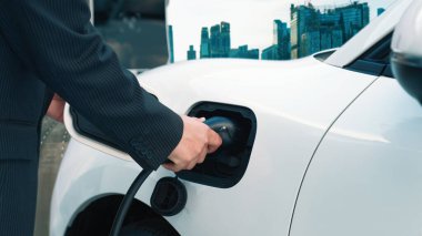 İlerici işadamı elektrikli aracını şarj kablosu ile şarj ediyor. Şarj istasyonundan şehrin arka planına bakıyor. Modern temiz enerji kampanyası konsepti için..