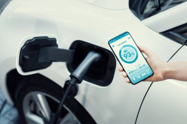 Battery status of electric vehicle displayed on smartphone application or software while vehicle is plugged into charging station for progressive future refueling. Battery status on phone screen.